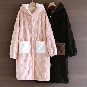 Pode incluir: Dois roupões de banho com capuz de pelúcia. Um é rosa claro com bolsos brancos, o outro é preto com bolsos castanhos. Ambos têm um padrão xadrez e fechos de botão. Os roupões estão pendurados em cabides de madeira.