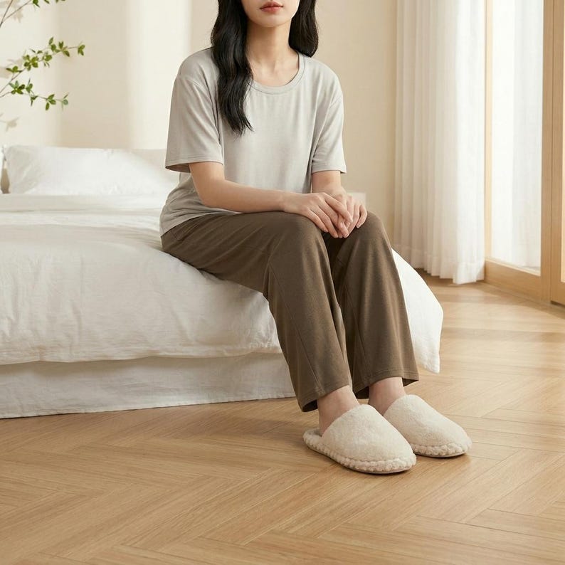 May include: A light gray short-sleeved crew neck t-shirt paired with dark brown lounge pants and cream-colored fuzzy slip-on slippers. The outfit consists of a soft-textured top and loose-fitting trousers, complemented by plush, textured footwear.