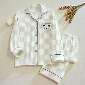 Könnte beinhalten: Ein hellgrünes und weiß kariertes Pyjama-Set. Das langärmlige Oberteil hat einen Panda-Bären-Aufnäher auf der Brusttasche. Das Set beinhaltet ein Hemd mit Knöpfen und eine passende Hose im Shorts-Stil. Schwarze Zierleisten akzentuieren Kragen, Manschetten und Tasche.