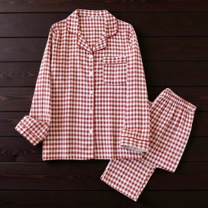 Könnte beinhalten: Ein rot-weiß kariertes Pyjama-Set. Das langärmlige Hemd hat eine Brusttasche und rote Paspeln. Die passende Hose ist ebenfalls kariert. Das Set wird auf einer dunklen Holzoberfläche präsentiert.