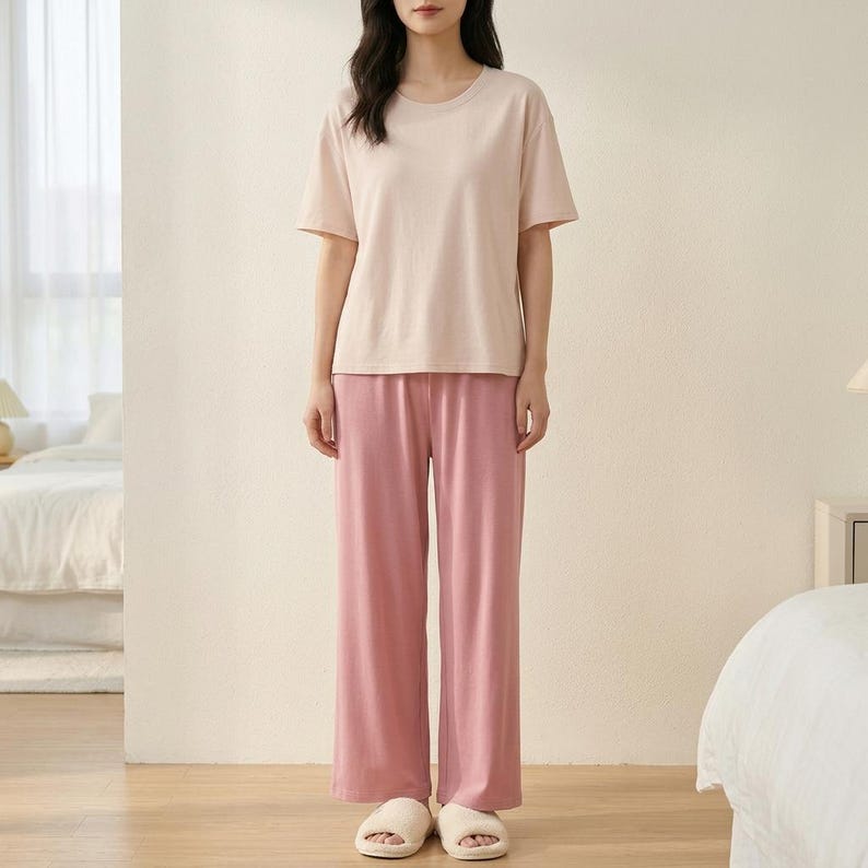 May include: A short-sleeved crew neck t-shirt in a light beige color paired with loose-fitting, wide-leg trousers in a solid dusty rose pink color. The outfit is completed with a pair of light-colored, open-toed slip-on house slippers.