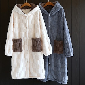 May include: Two plush hooded robes, one cream and one blue-grey, hang side-by-side. Each robe features a checkered pattern, brown pockets, and a brown-lined hood. The robes have button closures and are made of a soft, textured material.