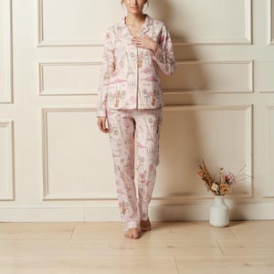 Könnte beinhalten: Rosa Pyjama-Set mit Pariser Motiv, einschließlich Illustrationen des Eiffelturms, Cafés und Figuren. Das Set besteht aus einem Hemd mit Kragen und Knöpfen sowie einer passenden Hose. Der Stoff wirkt weich und bequem, ideal zum Schlafen.