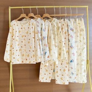 Puede incluir: Varios conjuntos de pijamas color crema cuelgan de una percha dorada. Los pijamas presentan una parte superior de manga larga y pantalones, ambos adornados con un patrón repetido de pequeños y coloridos personajes de dibujos animados. La tela parece ligera.