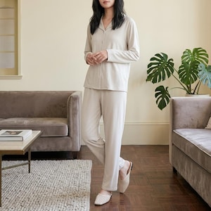 Könnte beinhalten: Beiges Pyjama-Set, bestehend aus einem langärmeligen Hemd mit Knöpfen und einer passenden Hose. Die weiche, neutrale Farbe der Nachtwäsche wird durch hellfarbene Hausschuhe ergänzt. Das Outfit ist auf Komfort und Entspannung ausgelegt.