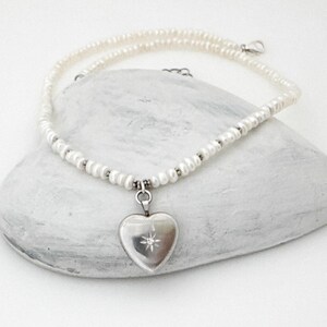 May include: A delicate necklace featuring a strand of small, white pearls. A silver heart-shaped locket with a starburst design hangs from the necklace. The necklace is displayed on a white, textured surface.