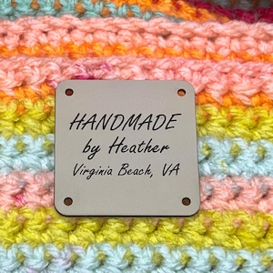 May include: Close-up of a handmade crochet item in vibrant colors including orange, pink, yellow, and blue. A tag reads "HANDMADE by Heather Virginia Beach, VA". The yarn is arranged in horizontal stripes.