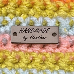 May include: Close-up of a handmade crochet item with horizontal stripes in shades of pink, yellow, orange, and blue. A small rectangular tag with the text "HANDMADE by Heather" is attached to the item.
