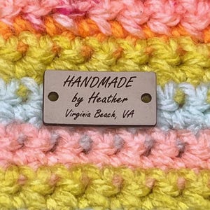 May include: Close-up of a handmade crochet item in a variety of colors, including orange, pink, yellow, and blue. The item has a textured appearance and features a small metal tag that reads "HANDMADE by Heather Virginia Beach, VA."