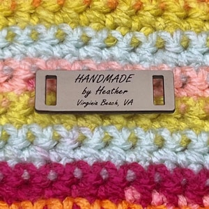May include: Close-up of a handmade crochet item with horizontal stripes in various colors, including orange, pink, yellow, and blue. A rectangular tag reads "HANDMADE by Heather Virginia Beach, VA."