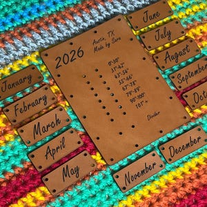 May include: A leather planner with the year 2026, temperature ranges, and month labels. The planner is surrounded by a colorful, striped crocheted blanket. The text "Austin, TX Made by Sara" is visible.