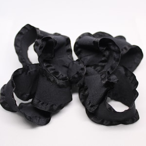 May include: A close-up of a black ribbon with a ruffled edge. The ribbon is made of a soft, silky material and is arranged in a loose, circular shape. The edges of the ribbon have a scalloped trim.