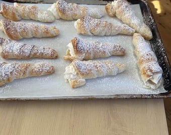 May include: A tray of freshly baked cream horns dusted with powdered sugar. The pastry shells are golden brown and filled with a white cream filling. The dessert is arranged on a sheet of parchment paper.