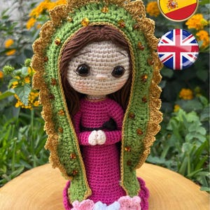 May include: A handmade crochet doll depicting a religious figure. The doll features a pink dress, green and gold accents, and a detailed face. The doll is approximately 6 inches tall and is displayed on a wooden surface.