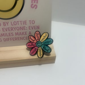 May include: A flower-shaped pin with petals in various colours, each bearing a positive word such as "kind" and "unique". The pin rests on a wooden base, with a backdrop that reads "Smiles make a difference".
