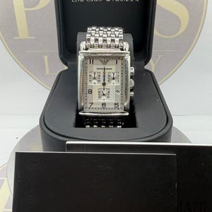 May include: A silver-toned Emporio Armani rectangular chronograph watch with a white dial, three sub-dials, and a metallic link bracelet. The watch is displayed inside a black presentation box with the Emporio Armani logo printed on the interior lid.