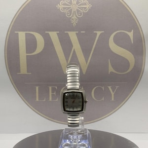 May include: A silver-toned wristwatch featuring a square-shaped face with a light-coloured dial, dark hour markers, and a flexible, segmented metal expansion band. The watch is displayed on a clear stand, showcasing its metallic finish and design.