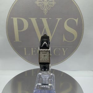 May include: A rectangular wristwatch with a silver case and a black leather strap. The watch face is light-coloured with silver hands and hour markers. The bezel is decorated with small, clear gemstones. The watch is displayed on a clear stand.
