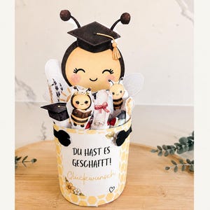 Peut inclure: Un seau décoratif avec un motif en nid d'abeille jaune et le texte allemand "DU HAST ES GESCHAFFT! Glückwunsch". Le seau contient des découpes en papier représentant une abeille avec une toque de diplômé, un diplôme et d'autres illustrations.