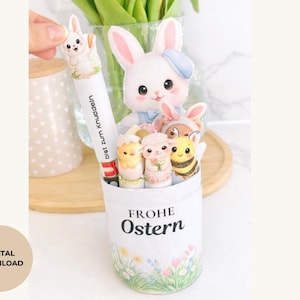 Op de afbeelding: Een witte container met de tekst "FROHE Ostern" en een bloemenontwerp, gevuld met paasfiguren, waaronder konijnen, een lam en een bij. Een pen met een konijnenopzetstuk wordt boven de container gehouden.