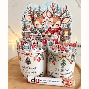 Könnte beinhalten: Zwei zylindrische Behälter mit weihnachtlichen Illustrationen und dem Text Advents kalender sowie 24 kleine Genussmomente. Sie enthalten nummerierte Stäbchen mit Festtagsmotiven. Ein horizontaler Stab vorne trägt die Aufschrift du bist mein Lieblingsmensch im Advent.