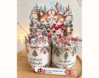 Peut inclure: Deux récipients cylindriques ornés d'illustrations de Noël avec le texte Advents kalender et 24 kleine Genussmomente. Ils contiennent des bâtonnets numérotés à motifs festifs. Un bâtonnet horizontal devant porte l'inscription du bist mein Lieblingsmensch im Advent.
