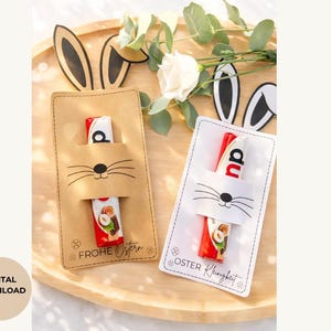 May include: Two Easter-themed gift card holders with bunny face cutouts, each holding a chocolate bar. One is tan with "FROHE OSTERN" text, the other white with "OSTER Kleinigkeit" text. The holders sit on a wooden tray with bunny ears and flowers.