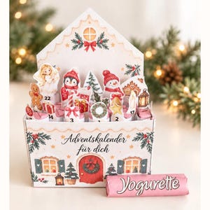 May include: A cardboard advent calendar shaped like a house with the text Adventskalender für dich, featuring illustrations of a gingerbread man, angel, penguin, snowman, and Christmas tree. A pink Yogurette chocolate bar sits in front.