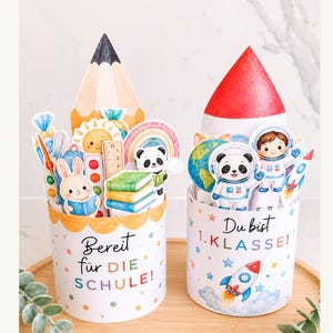 Peut inclure: Deux porte-crayons décoratifs sur le thème de l'école. L'un présente un dessus de crayon et le texte "Bereit für DIE SCHULE!" avec un lapin, des livres et un arc-en-ciel. L'autre a un dessus de fusée et le texte "Du bist 1. KLASSE!" avec des astronautes et un panda.