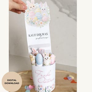 May include: An Easter-themed gift box with a floral design and the words "Frohe Ostern". The box contains decorative Easter eggs and bunny figures. The top of the box has a card with the text "KAUF DIR WAS schönes".