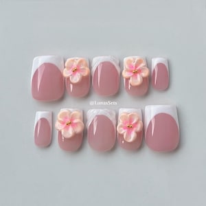 May include: A set of artificial nails with a French manicure design. The nails are pink with white tips and feature 3D flower embellishments. The flowers are light pink with gold centers. The text "@LunasSets" is visible.