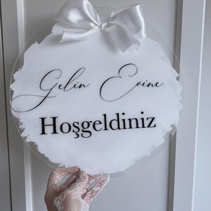 Könnte beinhalten: Runder, transparenter Schild mit den Worten "Gelin Evine Hoşgeldiniz" in schwarzer Schrift. Es hat einen weißen, pinselstrichartigen Hintergrund und ist mit einer weißen Satinschleife verziert.