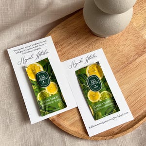 May include: Two white cards with text and a small green packet featuring lemon slices. The packet reads "EYUP SABRI TUNCER 1923 CESME LEMON REFRESHING TOWEL". The cards are on a wooden tray.