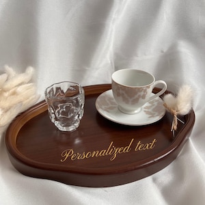 May include: A dark brown, oval wooden tray with a clear glass and a teacup and saucer. The teacup and saucer have a floral design. The tray has the words "Personalized text" in gold script.
