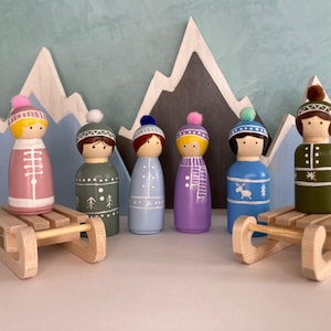 Op de afbeelding: Zes houten peg dolls in winterkleding, elk met een uniek hoed- en outfitontwerp, staan op kleine houten sleeën. De poppen zijn in verschillende kleuren, waaronder roze, groen, blauw en paars, met witte accenten. Een geschilderde bergachtergrond maakt de scène compleet.