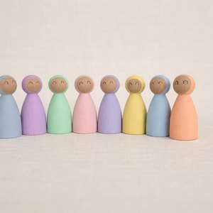 Puede incluir: Ocho muñecos de madera en colores pastel: azul claro, morado, verde, rosa, lavanda, amarillo, azul y melocotón. Cada muñeco tiene una cabeza de madera con una cara sonriente y un cuerpo con capucha.