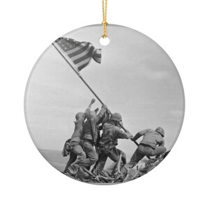 May include: A black and white ceramic ornament featuring the iconic image of the raising of the American flag on Iwo Jima. The round ornament has a gold string for hanging.