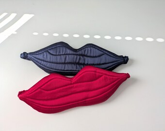 Op de afbeelding: Twee gewatteerde slaapmaskers in de vorm van lippen, één in diep marineblauwe satijnstof en de andere in felrode stof. Elk masker is voorzien van horizontale stiksels die de natuurlijke contouren van lippen nabootsen en heeft elastische zijbanden.