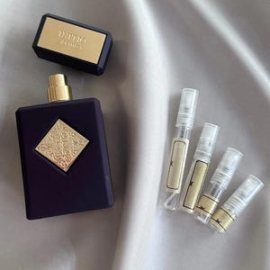 Pode incluir: Um frasco de perfume roxo escuro com detalhes dourados e uma tampa a condizer, juntamente com vários pequenos frascos de amostra. O frasco tem um emblema dourado e o nome da marca é visível na tampa. Sobre uma superfície sedosa cinza claro.