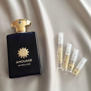 Pode incluir: Um frasco de perfume retangular azul marinho com uma tampa dourada e um emblema dourado, com a etiqueta "AMOUAGE INTERLUDE". Quatro pequenos frascos de amostra estão ao lado do frasco. O fundo é um tecido claro e sedoso.