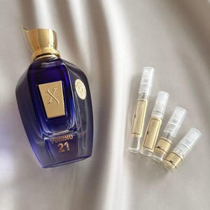 Puede incluir: Un frasco de perfume azul oscuro con tapa dorada y etiqueta dorada con la letra "X". El frasco está junto a cuatro pequeños viales de muestra con etiquetas doradas. El texto "TORINO 21" es visible en el frasco.