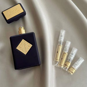 Pode incluir: Um frasco de perfume azul marinho com detalhes dourados e uma tampa a condizer. Vários pequenos frascos de amostra também estão presentes, todos sobre uma superfície acetinada. O texto "INITIO" é visível na tampa.