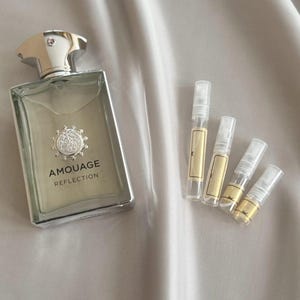 Pode incluir: Um frasco de perfume retangular com uma tampa prateada e o texto "AMOUAGE REFLECTION" está ao lado de quatro pequenos frascos de amostra. O frasco de perfume tem um corpo de vidro transparente com um líquido de cor clara no interior.