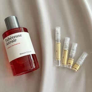 Pode incluir: Um frasco de perfume vermelho com tampa prateada e rótulo branco com o texto "Tubéreuse Astrale" e "extrait de parfum". Existem também quatro pequenos frascos de amostra de perfume com rótulos dourados.