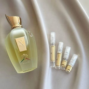 Pode incluir: Um frasco de perfume e vários frascos de amostra são exibidos em uma superfície de cetim. O frasco de perfume é verde claro com uma tampa dourada e um rótulo dourado. Os frascos de amostra têm detalhes e rótulos dourados.