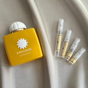 Pode incluir: Um frasco de perfume amarelo com uma tampa branca e o texto "AMOUAGE SUNSHINE". Vários frascos de amostra pequenos também estão presentes. Os itens estão dispostos em uma superfície texturizada de cor creme.