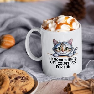 Könnte beinhalten: Weiße Keramik-Tasse mit einer Cartoon-Katzen-Illustration und dem Text "I knock things off counters for fun". Die Tasse ist mit Schlagsahne und Karamellsauce gefüllt. Ein Schokoladen-Chip-Keks befindet sich im Vordergrund.