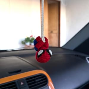 Puede incluir: Un adorno de coche de Spider-Man tejido a mano en rojo, azul y blanco, colgado de un cordón blanco. El salpicadero y las rejillas de ventilación del coche son visibles en el fondo. Un artículo decorativo.
