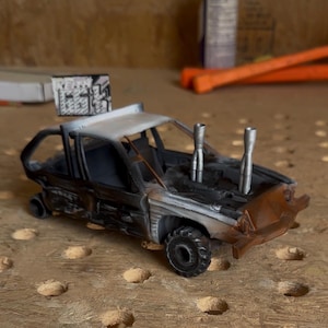 May include: A miniature model car featuring a weathered, distressed finish with shades of black, grey, and rust-colored paint. The vehicle includes two prominent silver exhaust pipes extending from the hood and a rectangular sign mounted on the roof.