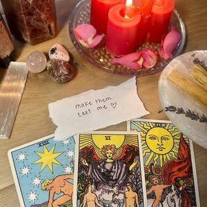 Puede incluir: Una colección de objetos espirituales sobre una superficie de madera. Incluye velas rojas encendidas, cartas del tarot, cristales, palos de palo santo y una nota escrita a mano que dice "make them text me". Las cartas del tarot presentan ilustraciones y las palabras "The Lovers" y "The Sun".